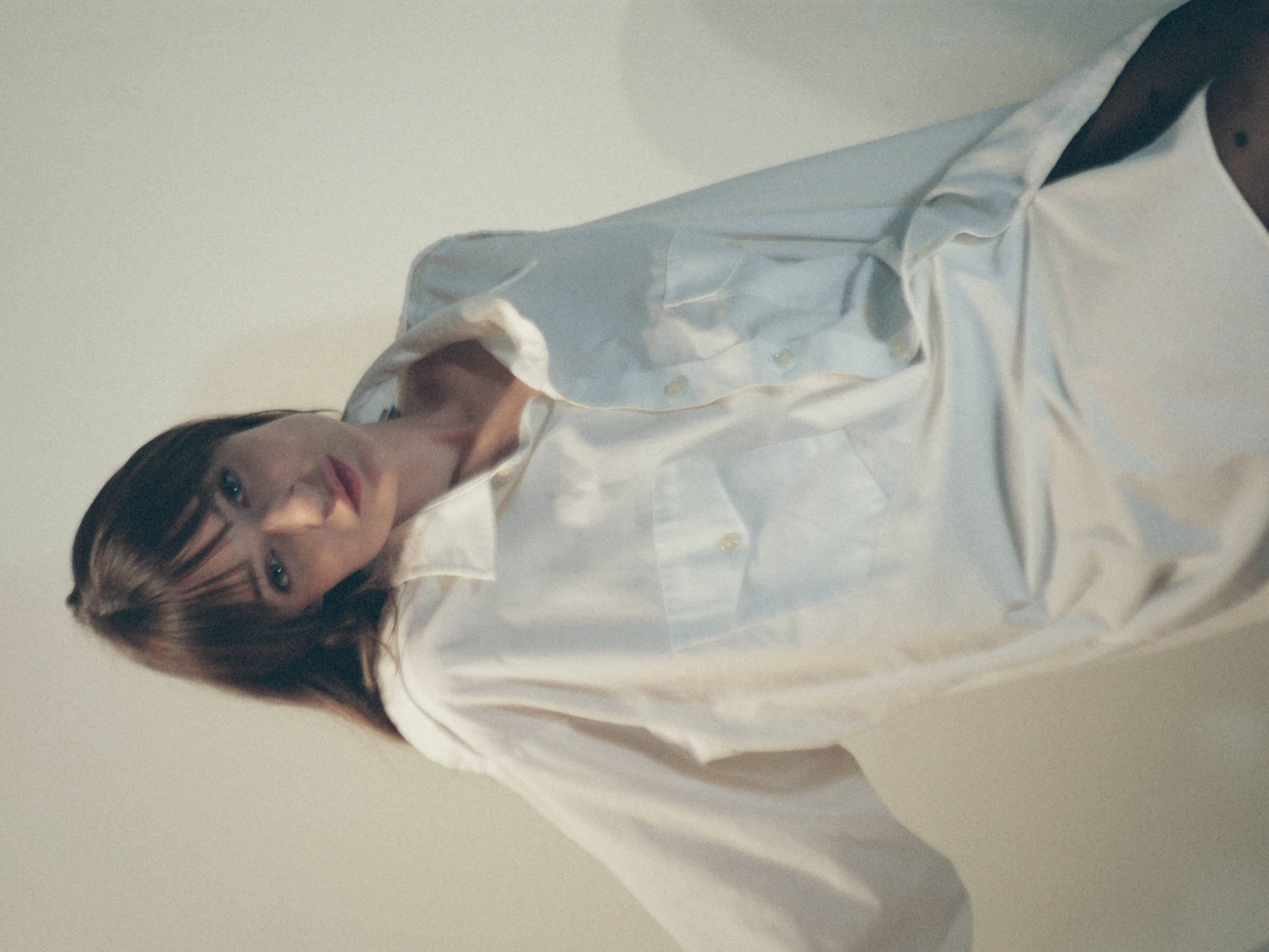 Kris Brouillette in an oversized white shirt, head tilted, one arm reaching out against a pale wall